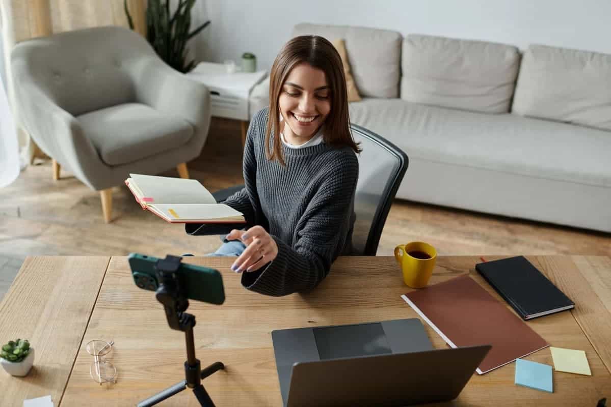 Mujer grabando un vídeo con el móvil mientras revisa su cuaderno de trabajo en casa